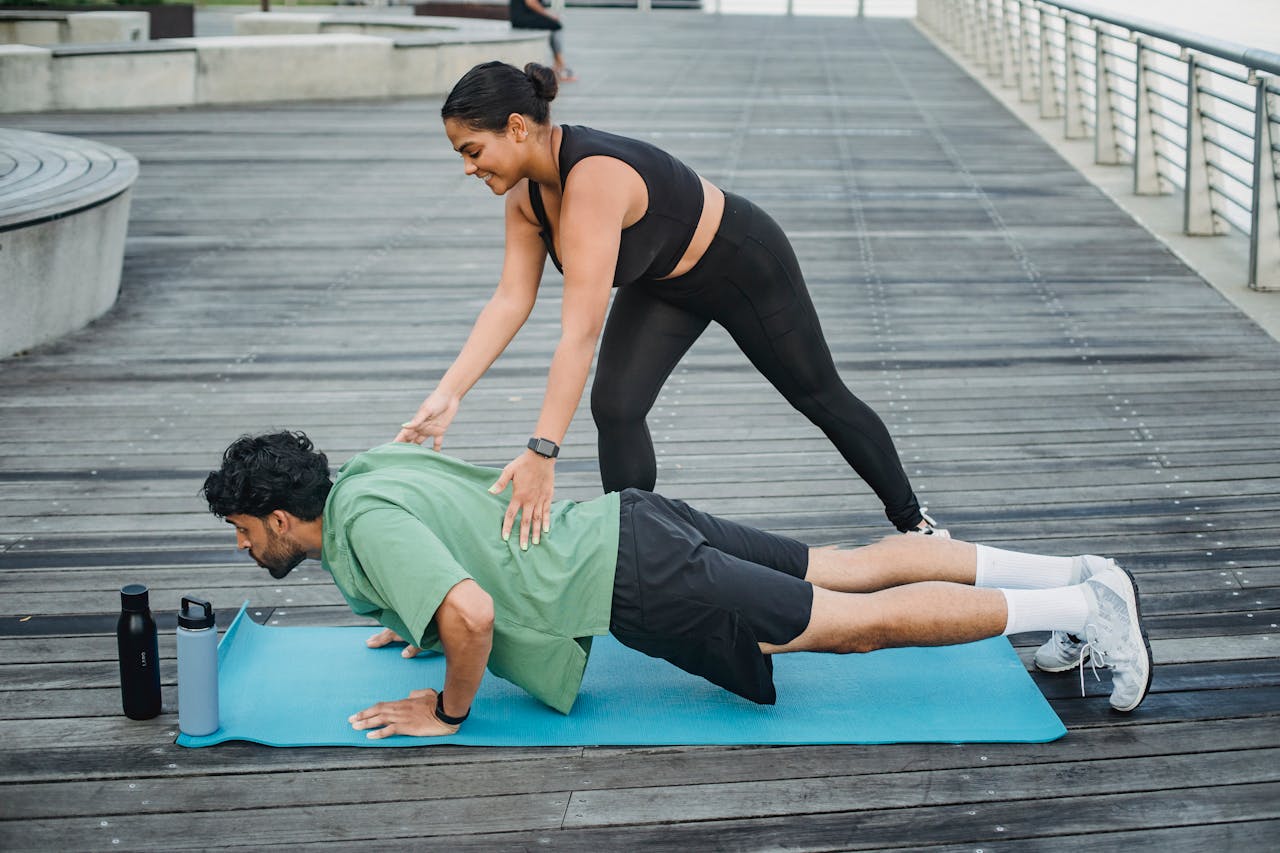 creative Man doing push-ups with assistance from a trainer on a boardwalk during summer.
